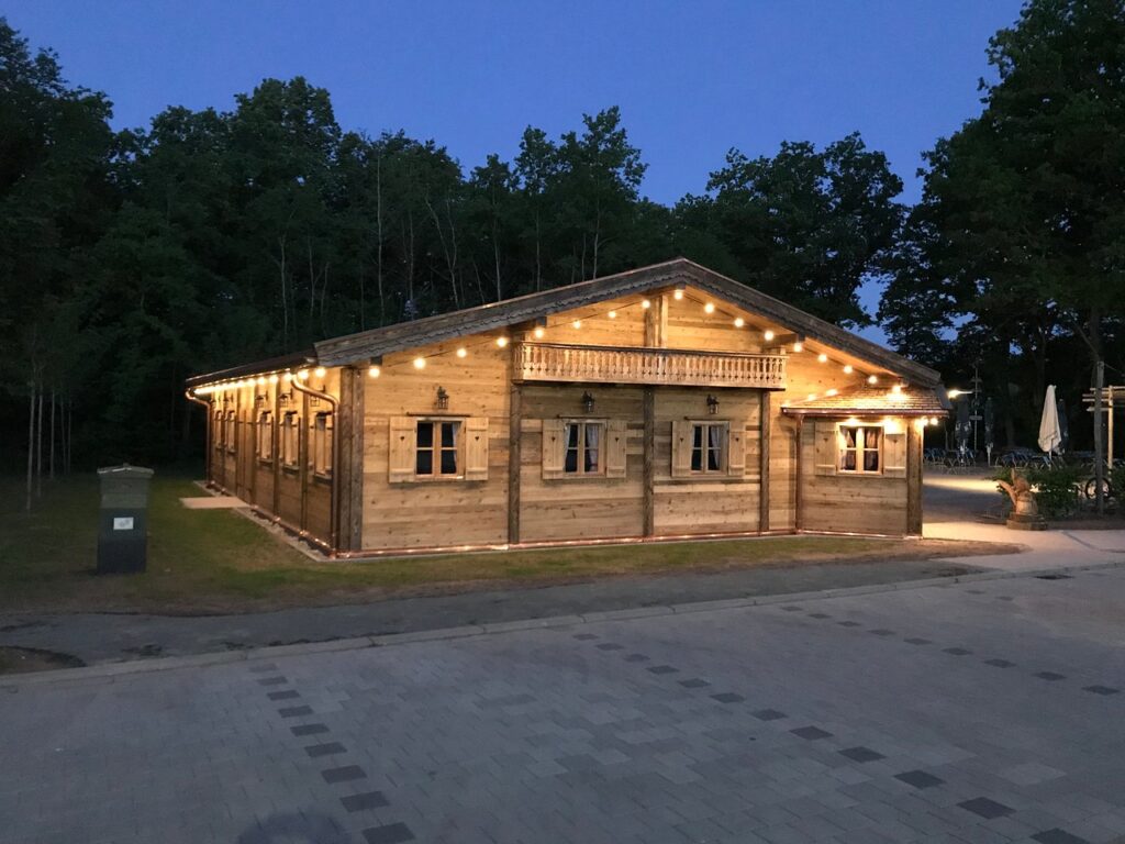 Holzhütte mit festlicher Beleuchtung, umgeben von Bäumen, bei Dämmerung. Pflasterweg im Vordergrund.