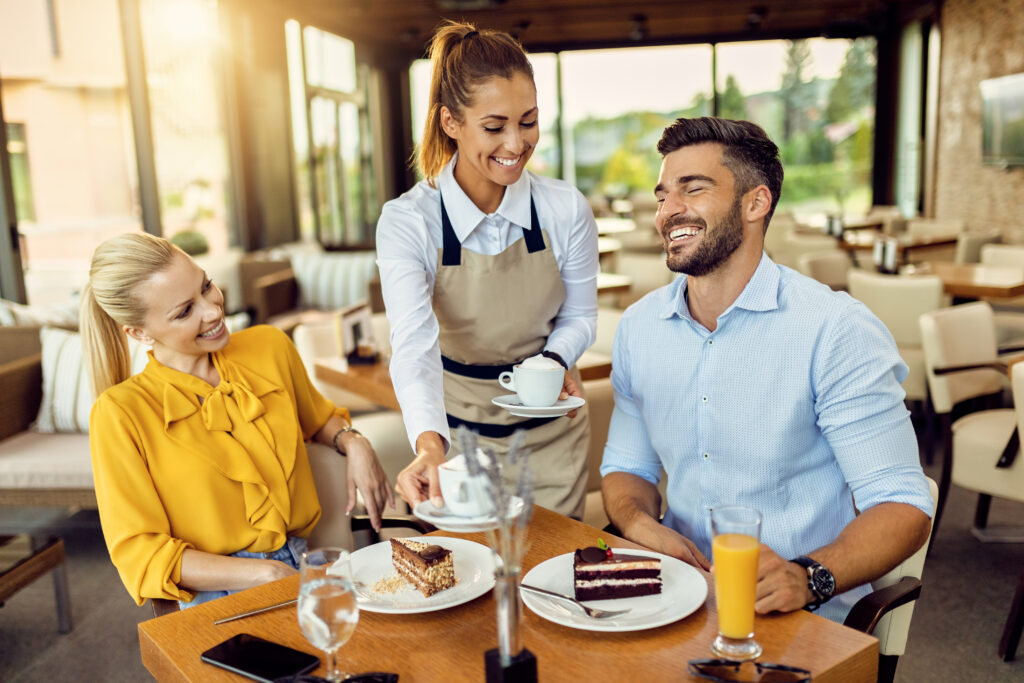 Eine blonde Frau und ein Mann lächeln, während eine Kellnerin ihnen Kaffee und Kuchen serviert.