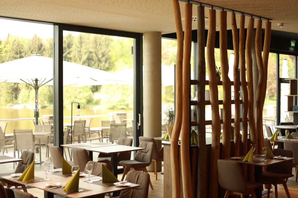 Helles Restaurant mit Holzelementen, Tischen mit gelben Servietten und Blick auf einen Außenbereich am Wasser.