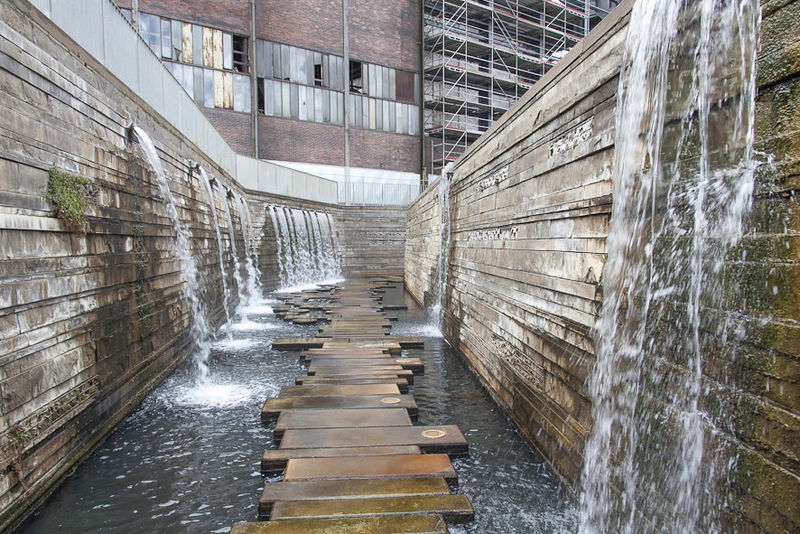 Futuristische Wasserkunst mit Holzstegen und Wasserfällen in einem urbanen Umfeld. Alte Industriegebäude im Hintergrund.