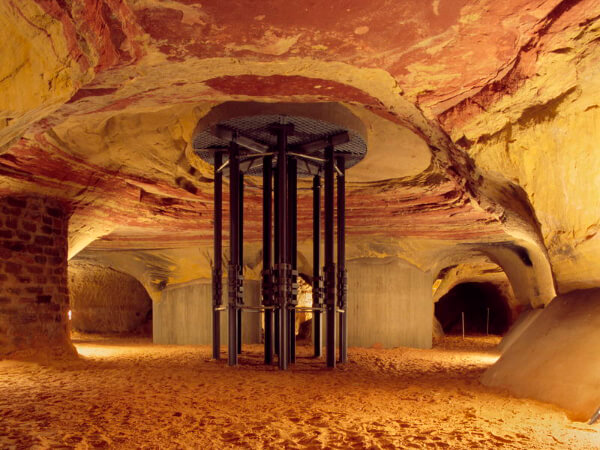 Eine unterirdische Höhle mit rotem Felsen, Sandboden und einer metallischen Struktur in der Mitte.