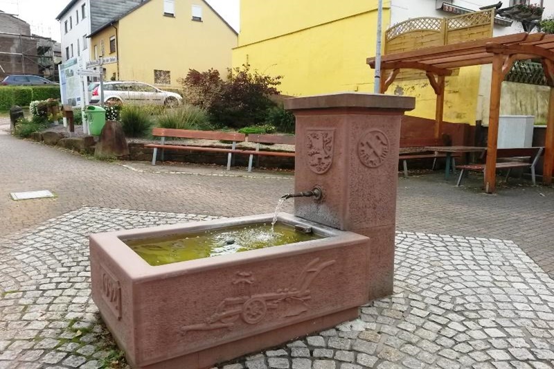 Brunnen mit Wasserlauf in einer gepflasterten Straße, umgeben von Bänken und farbigen Gebäuden.