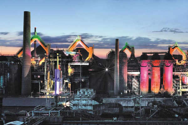 Industriegebiet bei Dämmerung mit bunten Lichtern und hohen Schornsteinen, die eine Stadtlandschaft beleuchten.