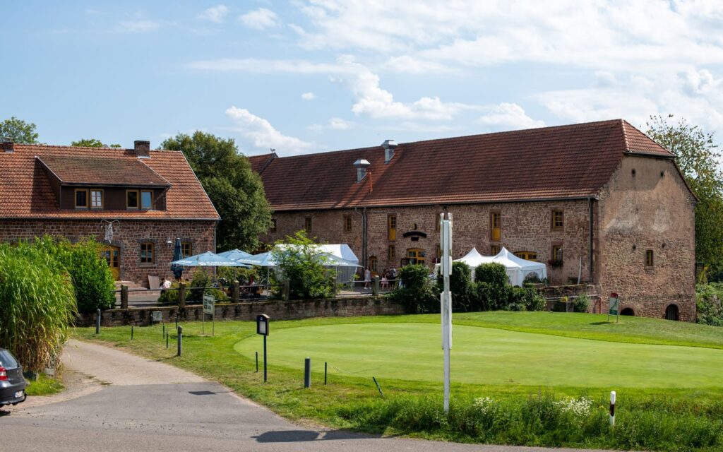 Ländliches Gebäude mit Terrassen, Zelten und einem Golfplatz im Vordergrund unter blauem Himmel.