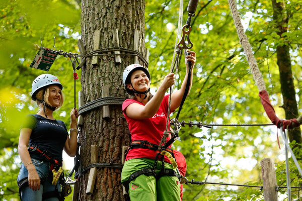 Zwei Frauen in Kletterausrüstung stehen an einem Baum, eine lächelt und hält eine Kletterausrüstung in der Hand.