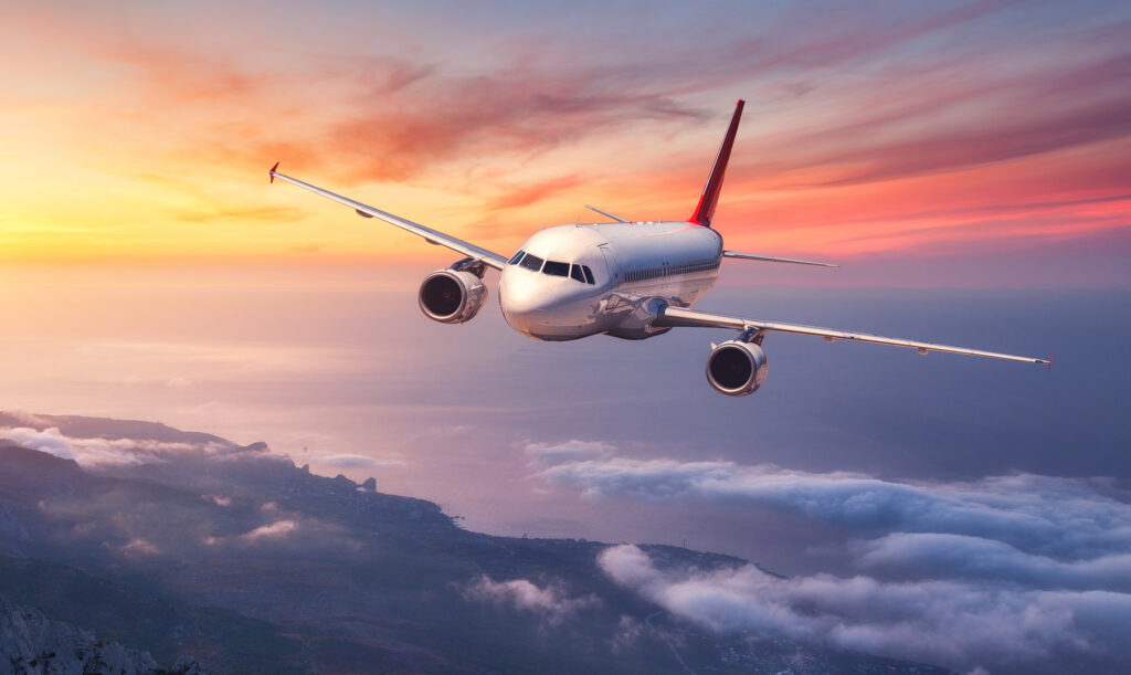 Ein Flugzeug fliegt über eine Küstenlandschaft bei Sonnenuntergang, umgeben von Wolken und sanften Farben des Himmels.